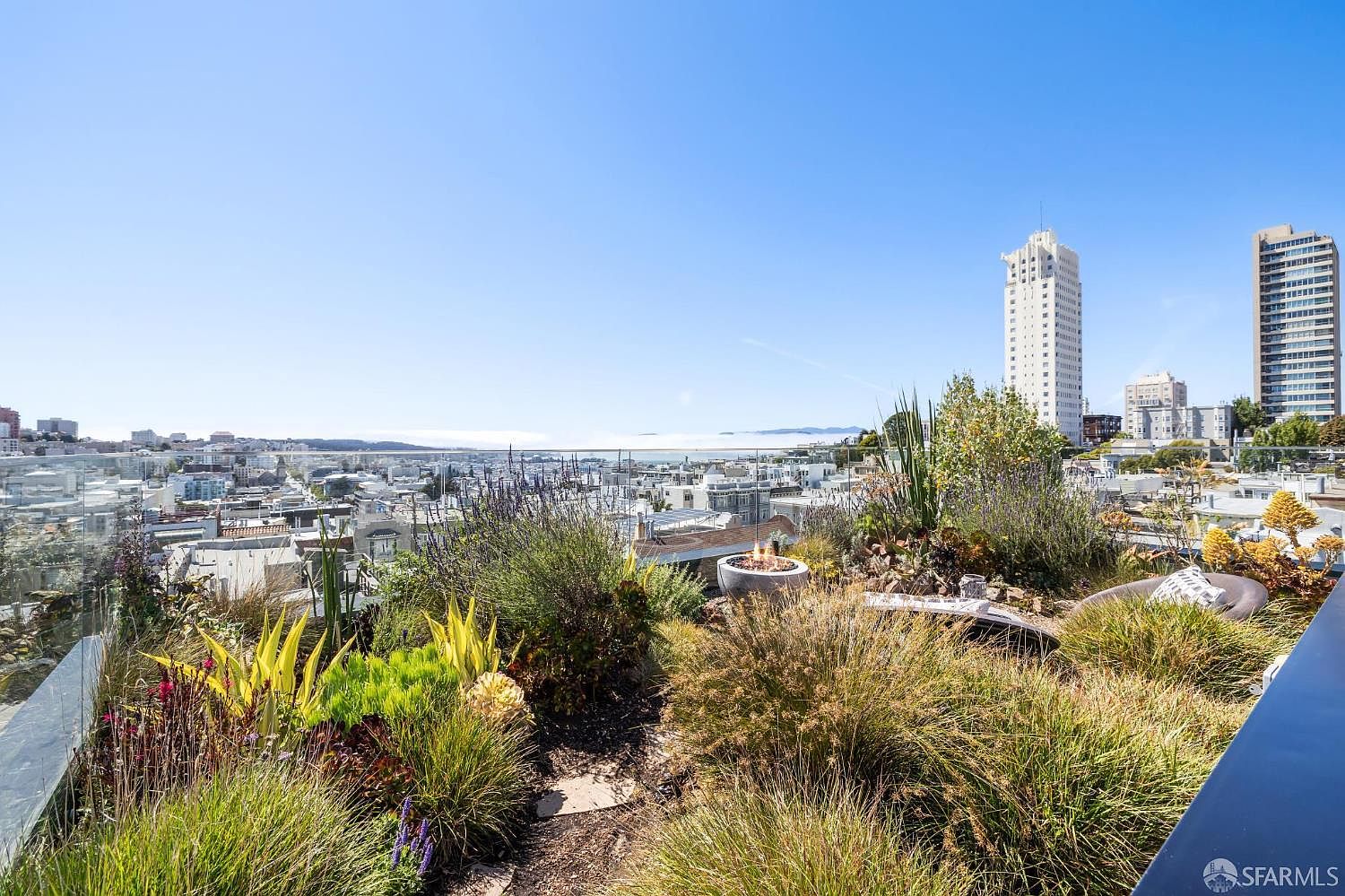 This elevated outdoor terrace offers a panoramic view of the city skyline under a clear blue sky. The space features a lush, landscaped garden with various grasses and succulents, centered around a circular fire pit. A glass railing provides an unobstructed perspective of the urban landscape, creating a sophisticated and serene atmosphere.