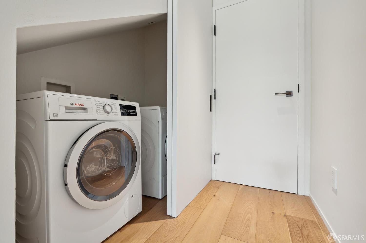 This laundry area features a white Bosch front-loading washing machine tucked into a recessed nook with light wood flooring. A matching dryer is positioned behind it, and a clean white door stands to the right, leading to another room. The space is minimalist and bright, emphasizing a functional and modern design.