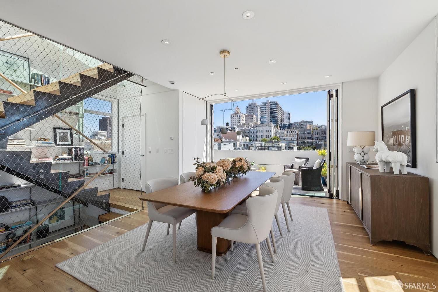 This bright and modern dining room features a central wooden table surrounded by six light-colored upholstered chairs, all set upon a textured area rug. To the left, a striking open-tread staircase is enclosed by a metal mesh railing, while a large glass wall opens to a balcony with city views. A wooden sideboard sits against the right wall, topped with a lamp and a decorative horse sculpture, creating a sophisticated and airy atmosphere.