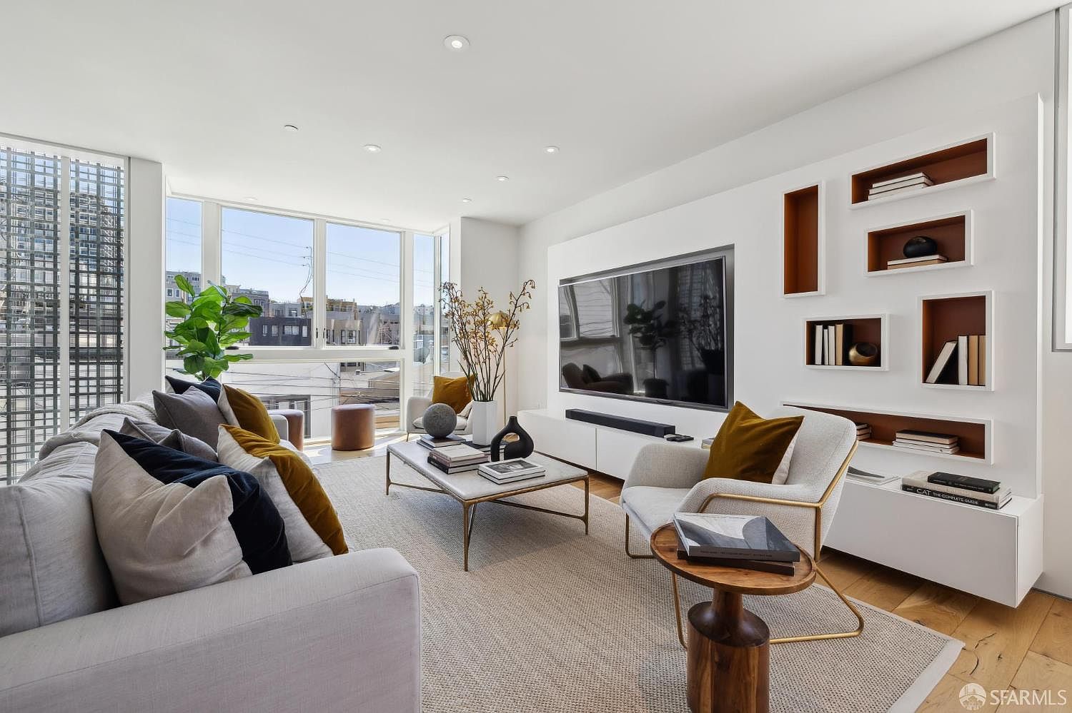 This bright and contemporary living room features a large, light-colored sofa accented with navy and mustard throw pillows, facing a wall-mounted television integrated into a custom white shelving unit with warm-toned recessed niches. A marble-topped coffee table sits on a textured area rug, complemented by a modern armchair and a small wooden side table. Large windows provide ample natural light and an urban view, creating an airy and sophisticated atmosphere.