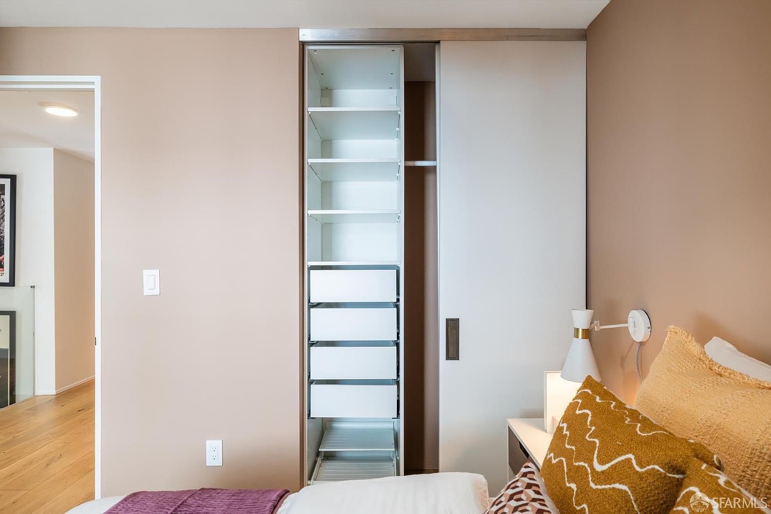 This bedroom features a modern, built-in sliding door closet with an open section revealing organized shelving and drawers. To the right, a portion of a bed with textured, mustard-colored pillows is visible, complemented by a sleek wall-mounted bedside lamp. The room is painted in a soft, neutral tone, creating a calm and contemporary atmosphere.