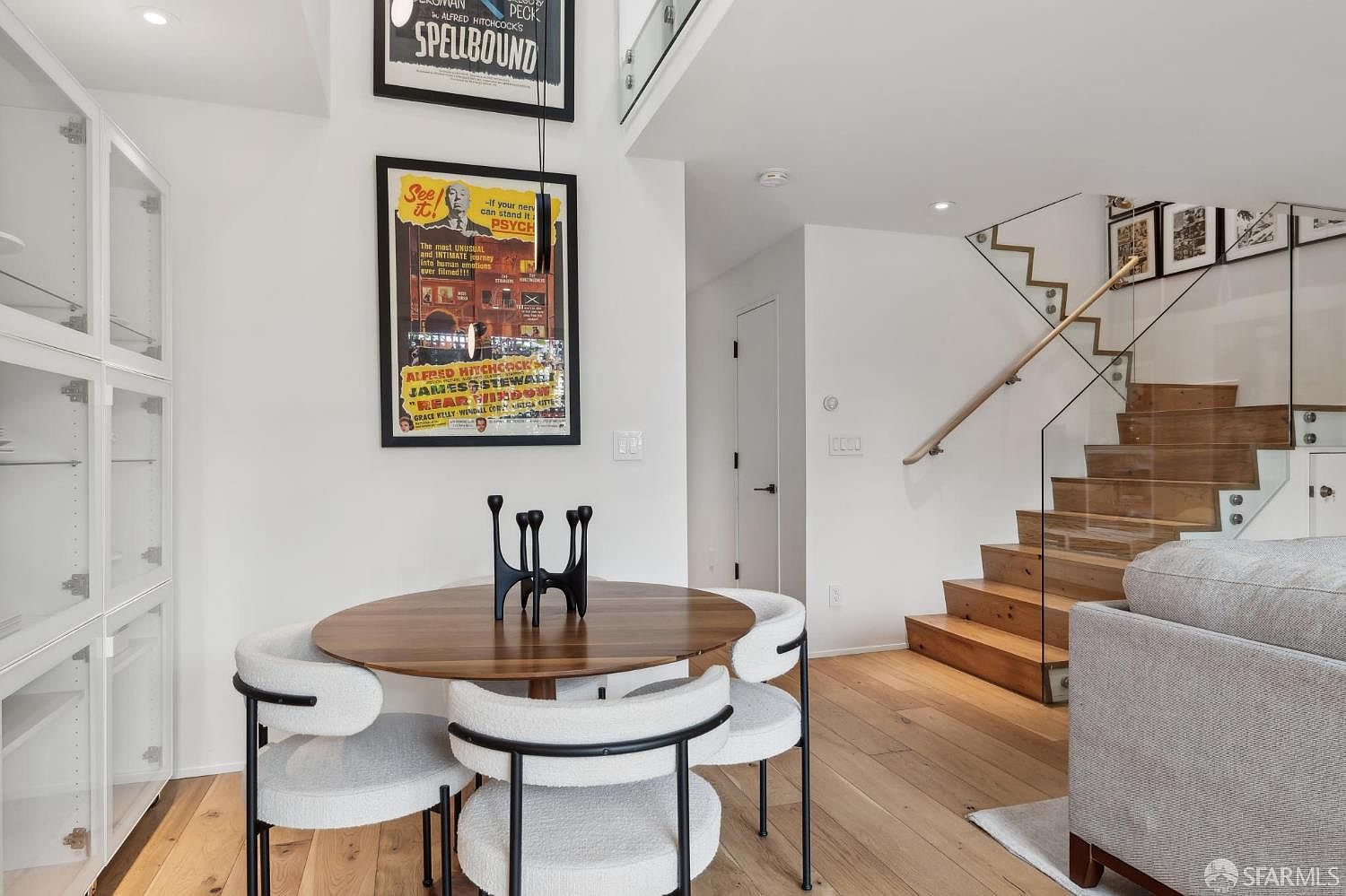 This contemporary dining area features a round wood-topped table surrounded by four modern chairs with light-colored upholstery and black metal frames. The space is accented by two framed vintage movie posters on the wall and a sculptural black centerpiece on the table. To the right, a staircase with wooden treads and a glass railing leads to an upper level, creating an open and airy atmosphere.