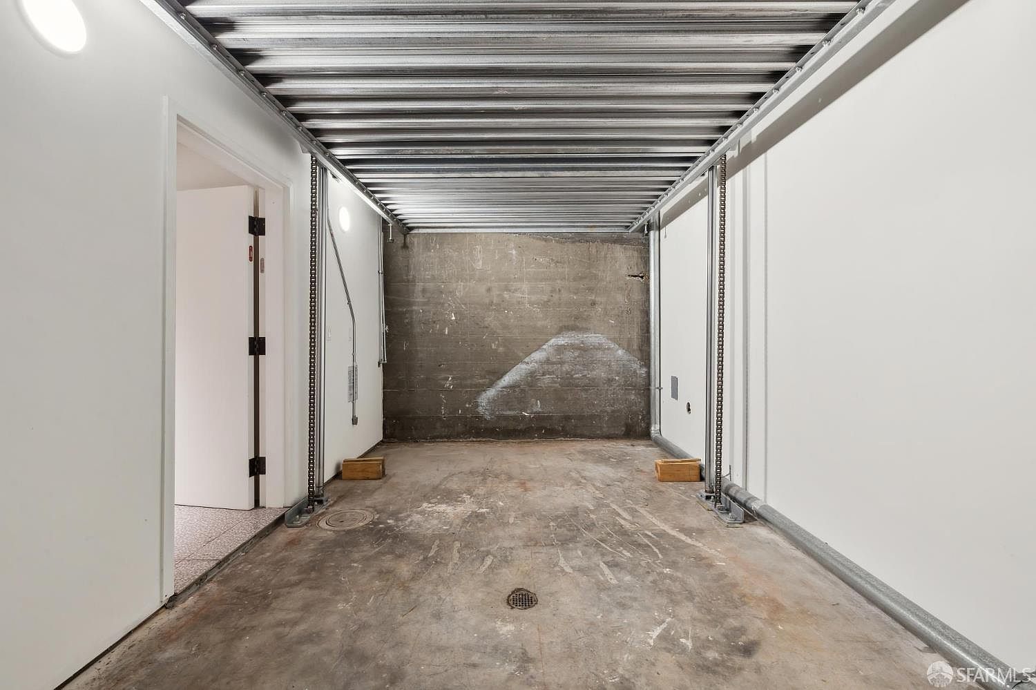 This image shows a utilitarian, industrial-style garage space with a concrete floor and a corrugated metal ceiling. The walls are finished in smooth white material, contrasting with the raw, textured concrete back wall, and the space includes a central floor drain and vertical metal support tracks. The perspective is a straight-on, eye-level shot looking into the depth of the structure, highlighting its functional and minimalist design.