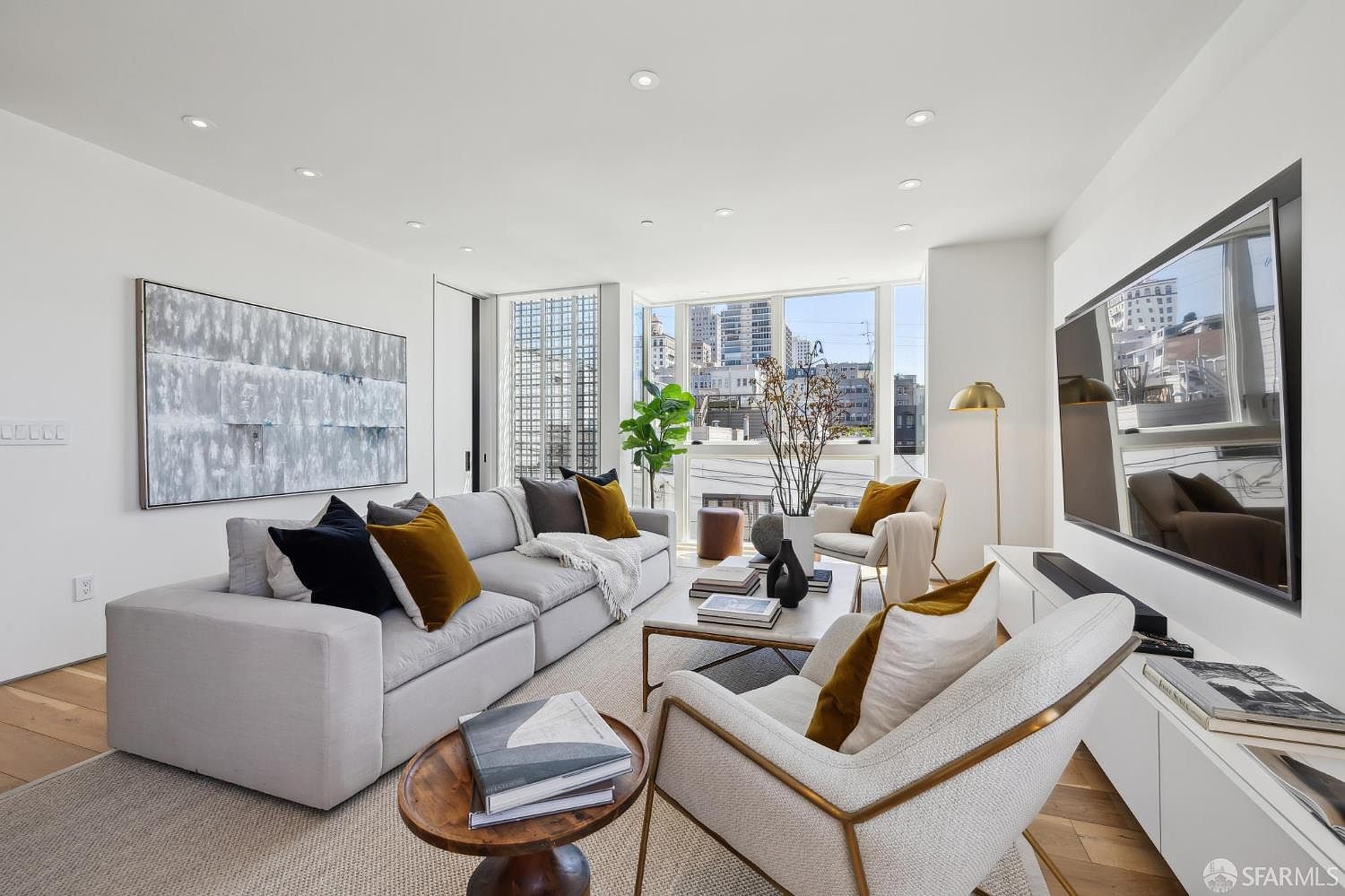 This bright and contemporary living room features a large, light-colored sectional sofa accented with dark and mustard-toned throw pillows. A central coffee table sits on a textured area rug, complemented by a pair of modern armchairs and a small wooden side table in the foreground. Large windows provide ample natural light and an urban view, while a large television is mounted on the wall above a sleek, low-profile media console.
