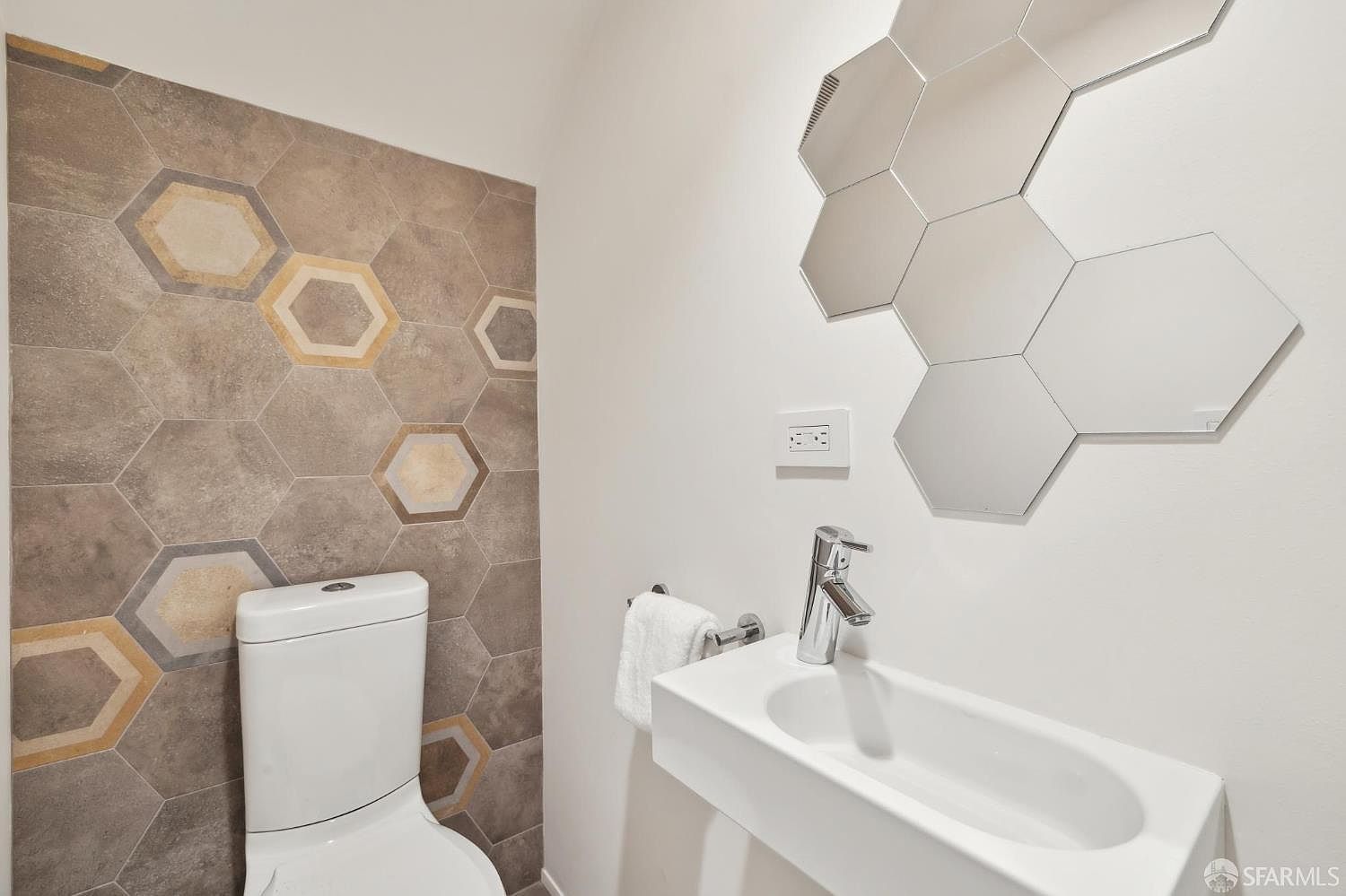 This modern powder room features a striking accent wall adorned with hexagonal tiles in shades of gray, white, and tan. A wall-mounted white vanity with a sleek chrome faucet sits beside a white toilet, while a decorative arrangement of hexagonal mirrors adds a contemporary touch above the sink. The space is bright and minimalist, utilizing geometric shapes to create a stylish and cohesive aesthetic.