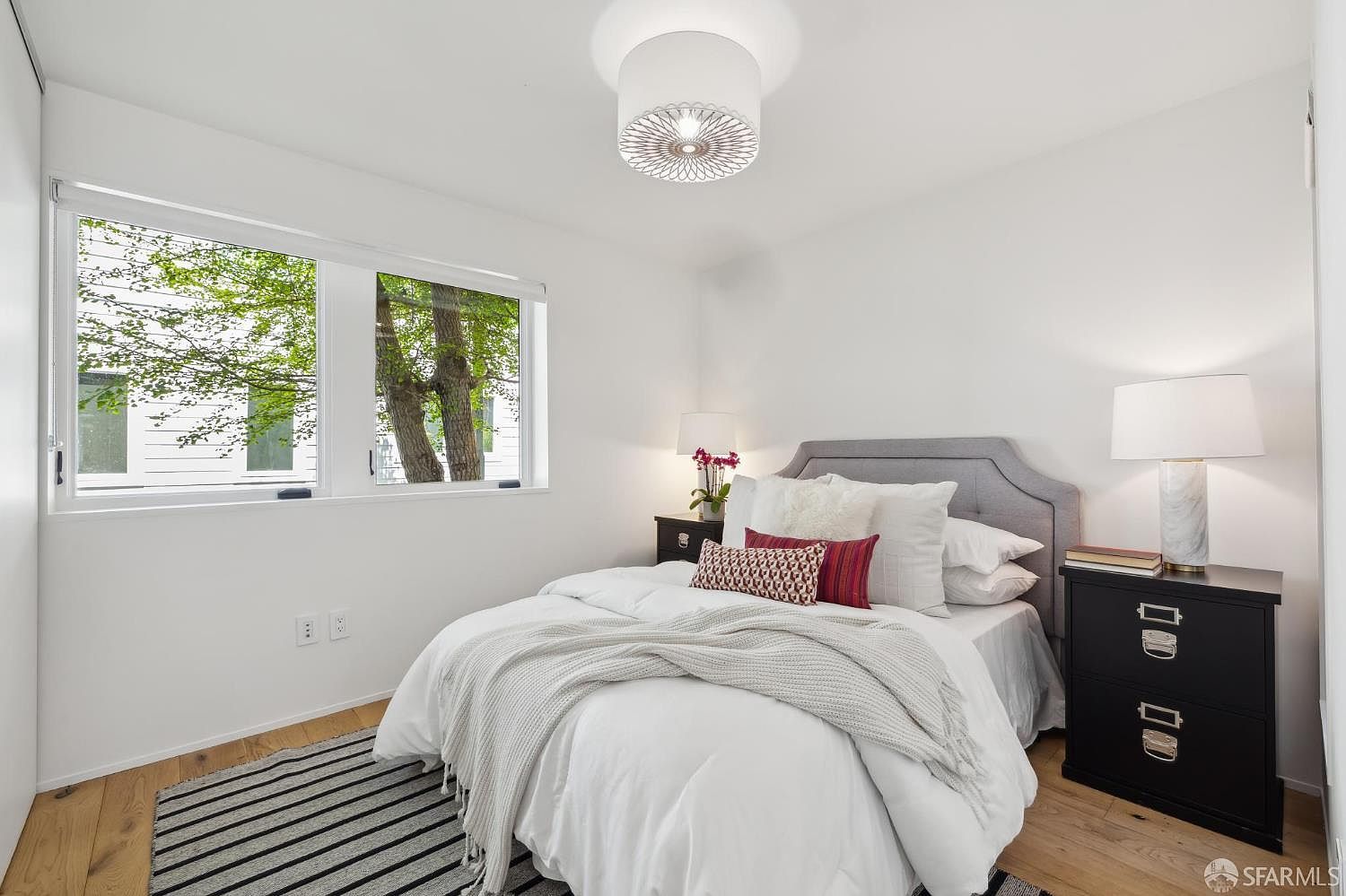 This bright and inviting bedroom features a neutral color palette with a light gray upholstered headboard and a crisp white bed ensemble. The room is accented by a patterned throw pillow, a textured throw blanket, and a striped area rug, all set against light wood flooring. Large windows allow natural light to fill the space, offering a view of mature trees outside.