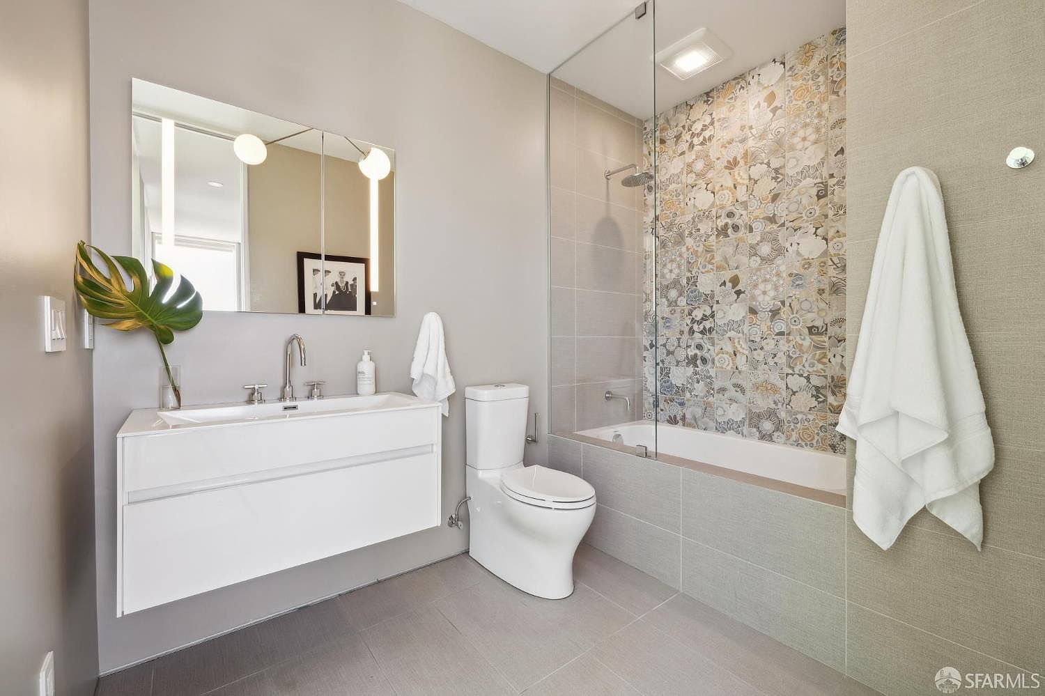 This modern bathroom features a floating vanity with a sleek sink, a wall-mounted toilet, and a bathtub enclosed by a glass partition. The shower area is accented with a decorative, patterned tile wall that contrasts with the neutral, large-format floor and wall tiles. The space is bright and contemporary, with a large mirror and minimalist lighting fixtures creating a clean, airy aesthetic.
