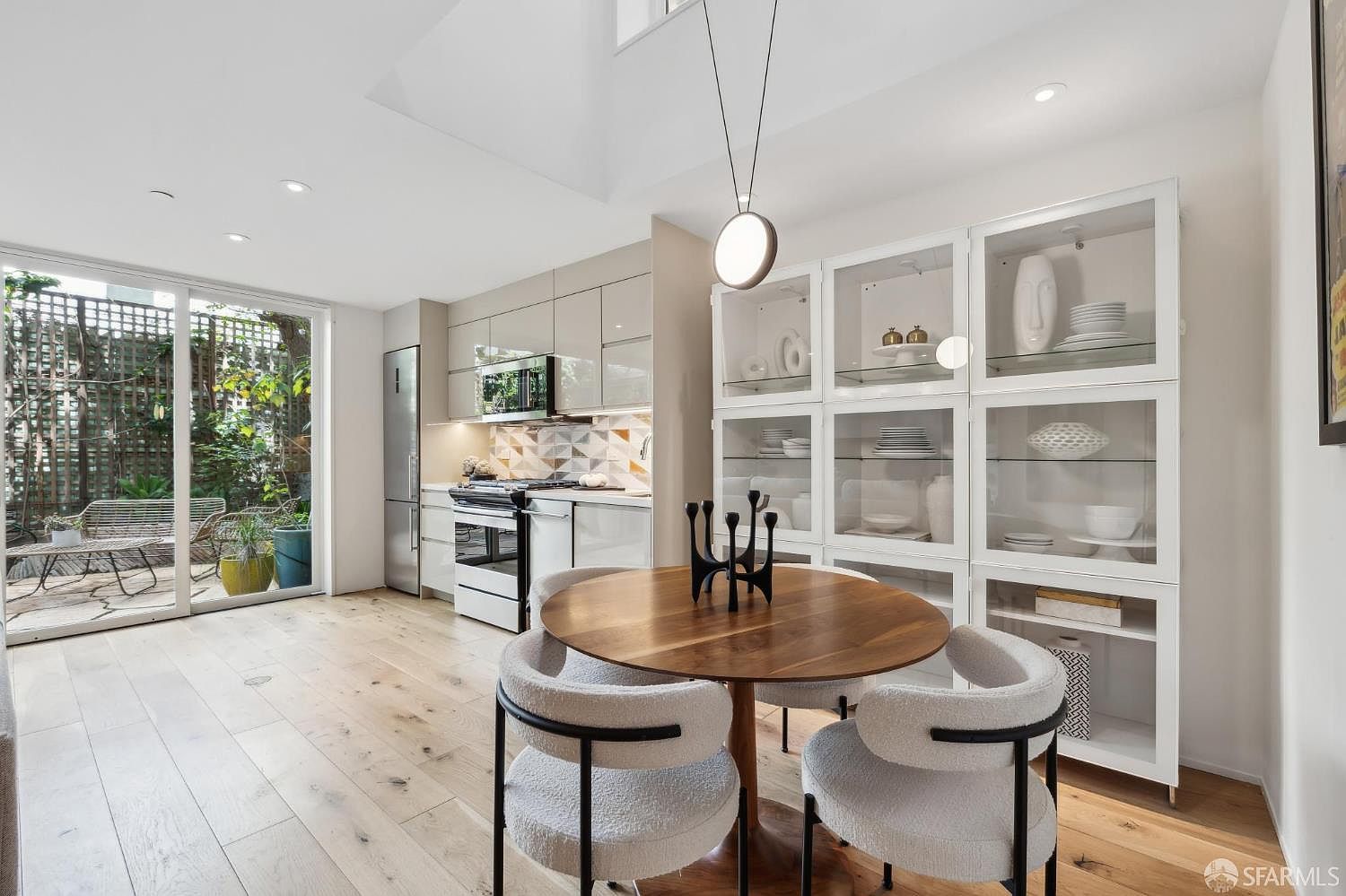 This bright, open-concept dining area features a round wooden table paired with four modern, textured chairs and a sculptural black centerpiece. To the right, a large white glass-front cabinet displays decorative dishware, while the adjacent kitchen showcases light-toned cabinetry and a geometric patterned backsplash. A sliding glass door provides a seamless transition to an outdoor patio, filling the space with natural light and creating an airy, contemporary atmosphere.