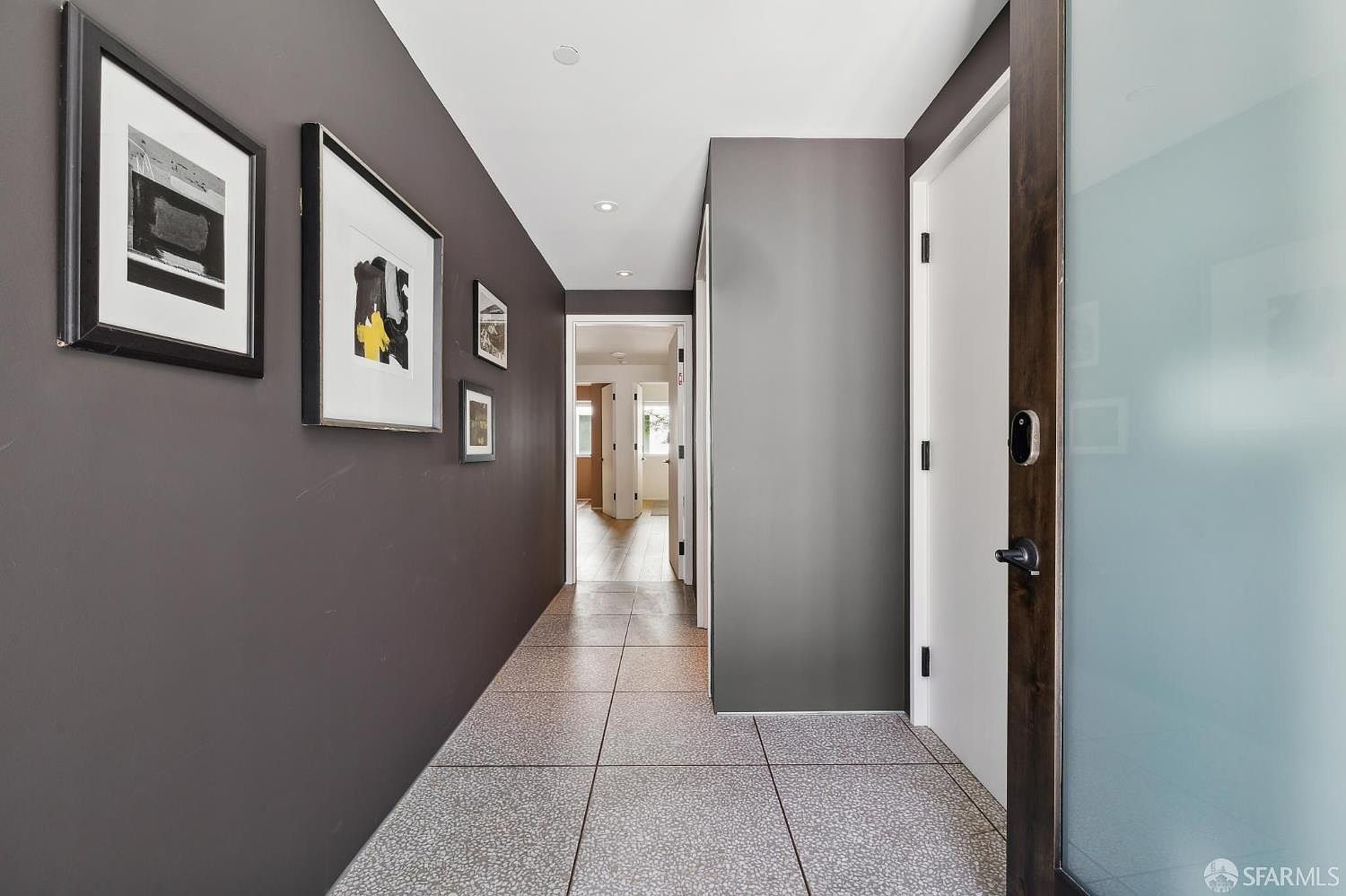 This modern hallway features a striking contrast between dark charcoal walls and a light-toned ceiling, creating a sophisticated and dramatic atmosphere. The floor is covered in large, speckled tiles, and the wall is adorned with a gallery of framed artwork. A doorway on the right leads to a room with a frosted glass door, while the hallway extends forward into a bright, open living space with light wood flooring.