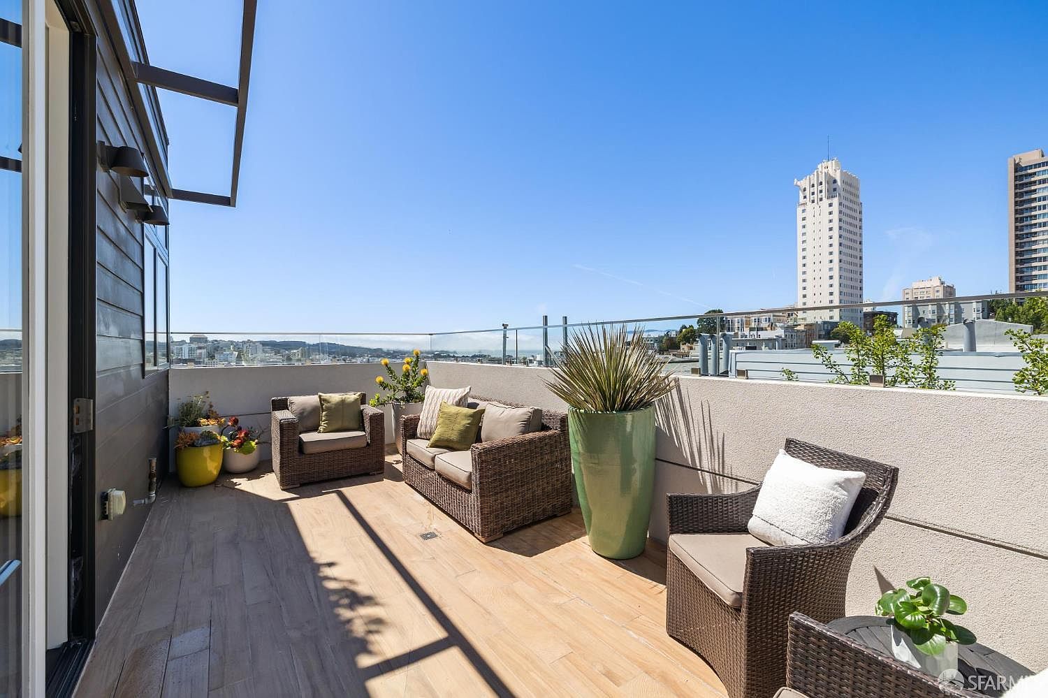 This spacious rooftop balcony features light-toned wood-look decking and a modern glass railing that provides an unobstructed view of the city skyline. The area is furnished with comfortable wicker-style outdoor seating, including a sofa and armchairs, accented by potted plants and a large decorative planter. The perspective is a wide-angle shot from the doorway, capturing the inviting atmosphere and the bright, sunny urban setting.