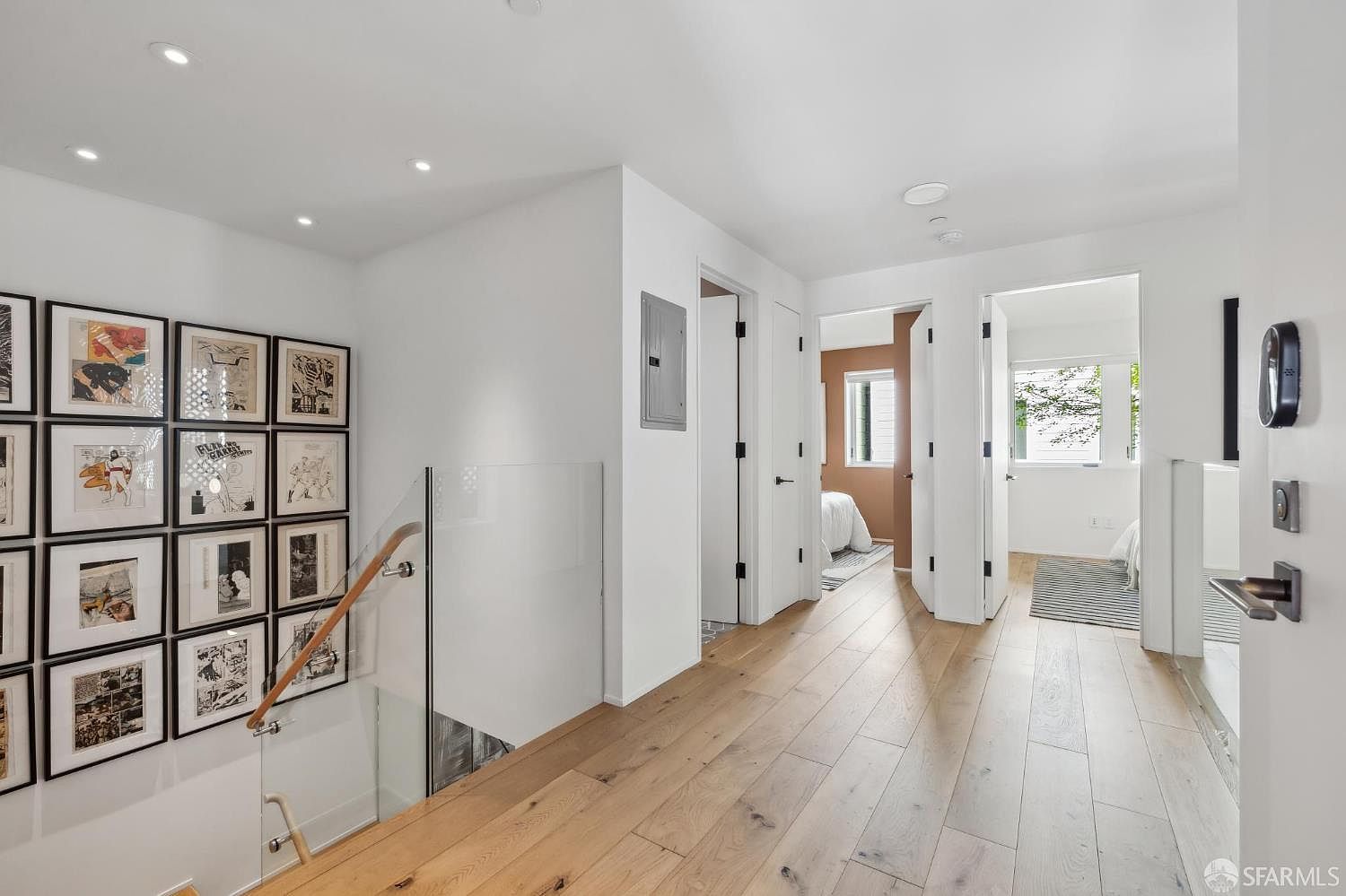 This bright, modern hallway features light wood flooring and a clean, minimalist aesthetic. A glass-railed staircase leads to a lower level, while a gallery wall of framed artwork adds character to the space. The hallway provides access to multiple rooms, including a bedroom visible in the background, and is illuminated by recessed ceiling lights.