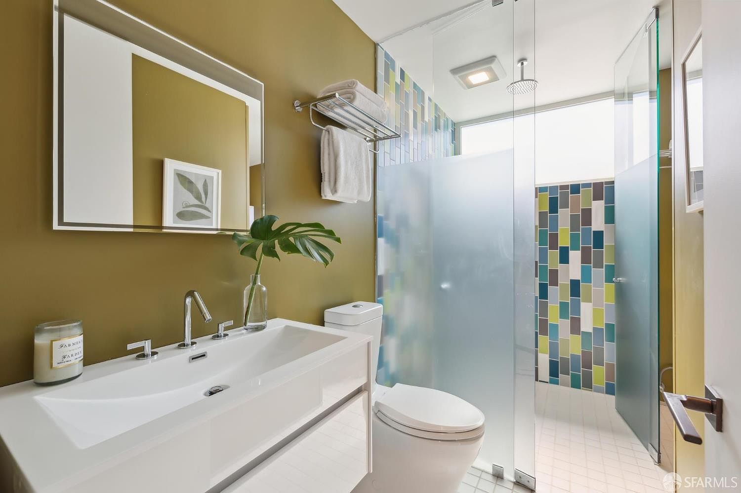 This modern bathroom features a sleek, wall-mounted white vanity with a rectangular sink and a large, frameless mirror. The space is accented by a bold olive-green wall and a walk-in shower enclosed by frosted glass, which showcases a vibrant, multi-colored mosaic tile wall. The perspective is a straight-on eye-level shot that highlights the clean lines and contemporary design of the fixtures.