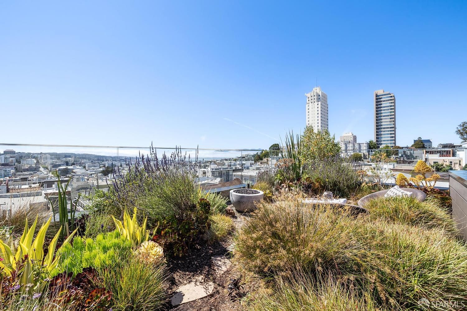 This rooftop terrace features a lush, landscaped garden with a variety of grasses, succulents, and lavender, creating a serene urban oasis. A modern, circular fire pit sits nestled among the greenery, while a glass railing provides an unobstructed, panoramic view of the city skyline and distant hills. The perspective is from a low angle within the garden, emphasizing the contrast between the natural textures of the plants and the architectural density of the city beyond.