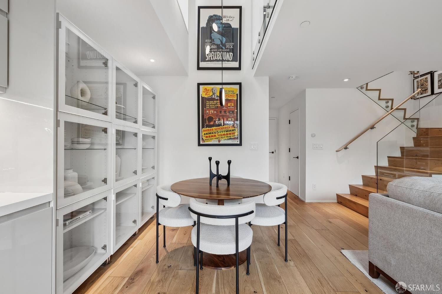 This bright dining area features a round wooden table surrounded by four modern chairs with white upholstered seats and black metal frames. To the left, a large white display cabinet with glass doors provides ample storage, while the wall is decorated with two framed vintage movie posters. A wooden staircase with a glass railing is visible on the right, leading to an upper level, creating an open and airy atmosphere.