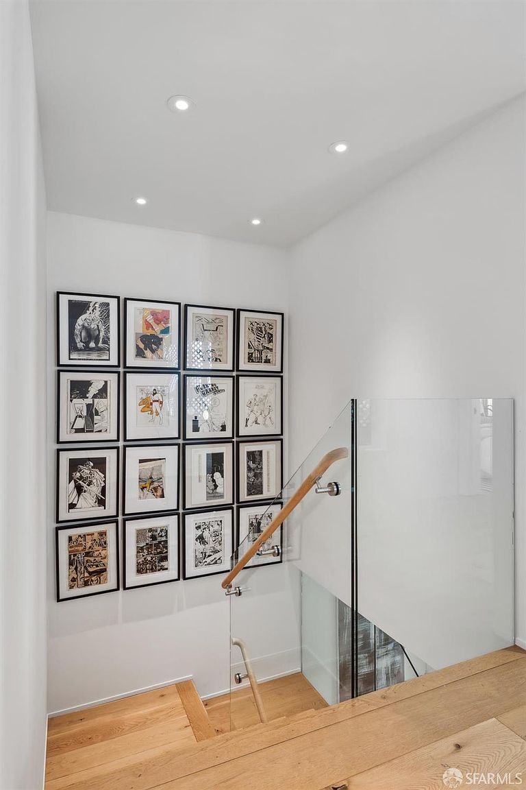 This interior view captures a modern staircase featuring light wood treads and a sleek glass railing with a minimalist wooden handrail. A prominent gallery wall displays a grid of sixteen framed black-and-white comic book panels, adding a unique artistic touch to the bright, white-walled space. The perspective looks down the staircase, highlighting the clean lines and contemporary architectural design of the home.