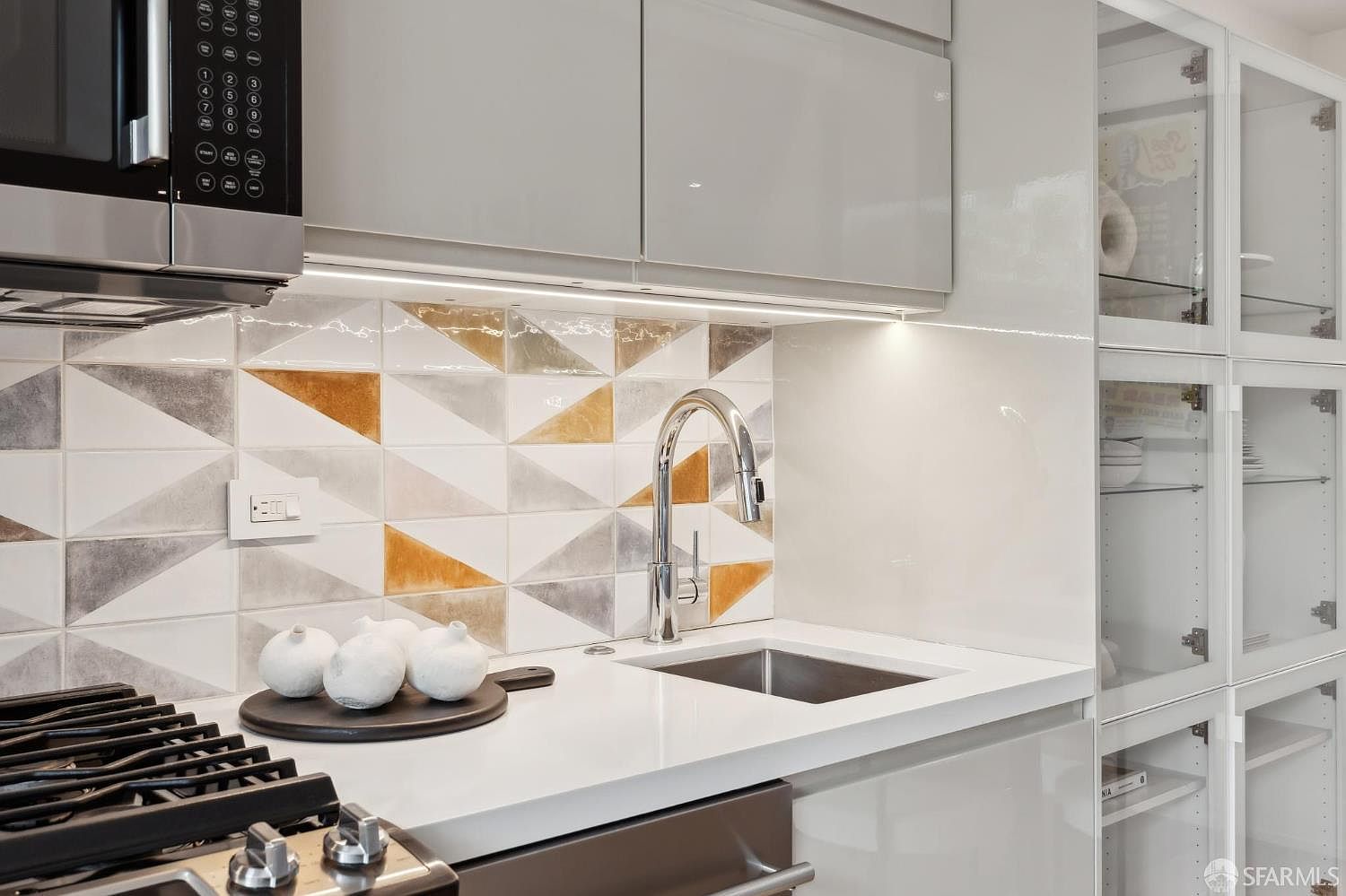 This modern kitchen area features sleek, high-gloss cabinetry and a striking geometric backsplash with metallic and white triangular tiles. A stainless steel faucet sits above a compact undermount sink, complemented by a white countertop and a black circular tray holding decorative white spheres. The space is illuminated by under-cabinet lighting, highlighting the contemporary design and clean lines of the workspace.