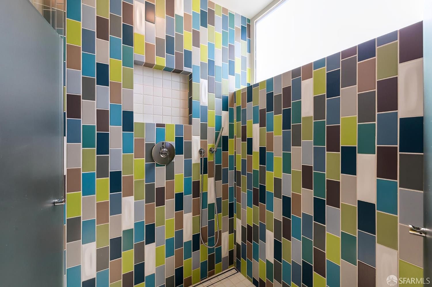This modern shower space features a vibrant, multi-colored mosaic tile pattern in shades of blue, green, and grey, creating a bold and artistic aesthetic. The shower includes a built-in recessed niche for storage, a sleek chrome shower head, and a handheld sprayer attachment. A high, horizontal window allows natural light to brighten the space, while the clean lines and contemporary fixtures provide a polished, high-end feel.