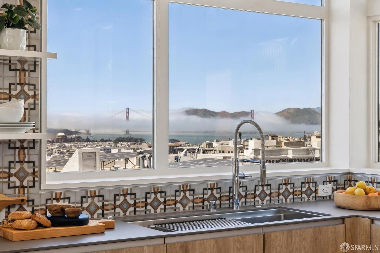 This kitchen features a sleek, modern design with light wood cabinetry and a neutral countertop, centered around a stainless steel sink and faucet. A striking patterned tile backsplash adds visual interest, while large windows provide a breathtaking, panoramic view of the Golden Gate Bridge partially shrouded in fog. The perspective is from the sink looking out, emphasizing the seamless connection between the refined interior space and the iconic San Francisco landscape.
