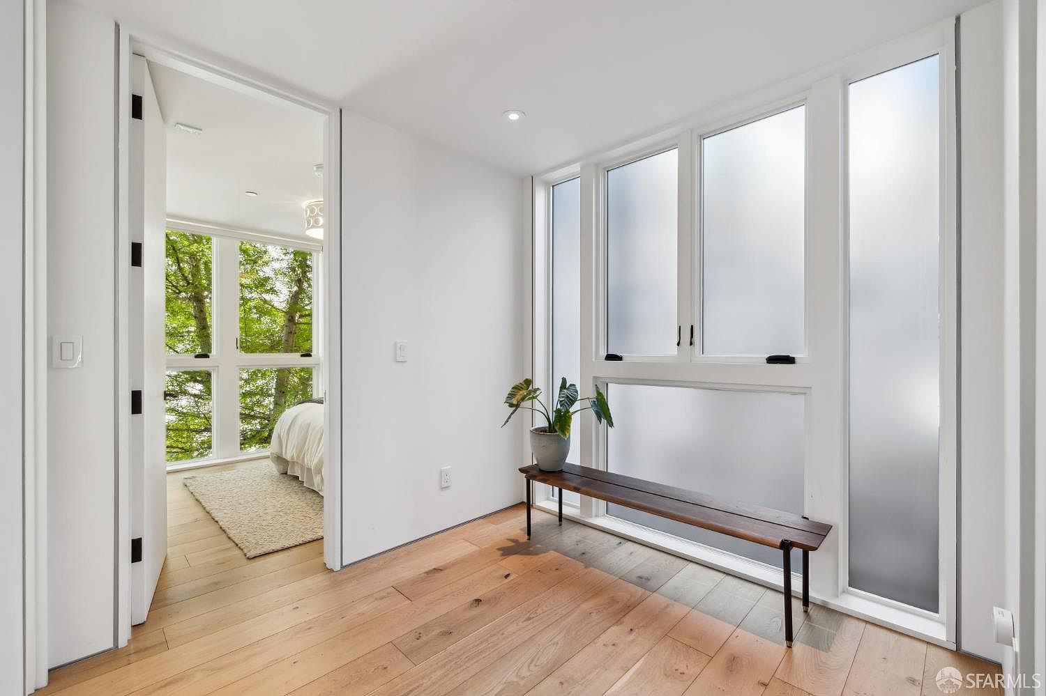 This bright, minimalist hallway features light wood flooring and crisp white walls, creating an airy and modern atmosphere. A long, dark wood bench sits beneath a series of large, frosted windows that allow soft, diffused light to fill the space. Through an open doorway, a glimpse of a bedroom with large windows overlooking lush greenery adds a sense of depth and connection to the outdoors.