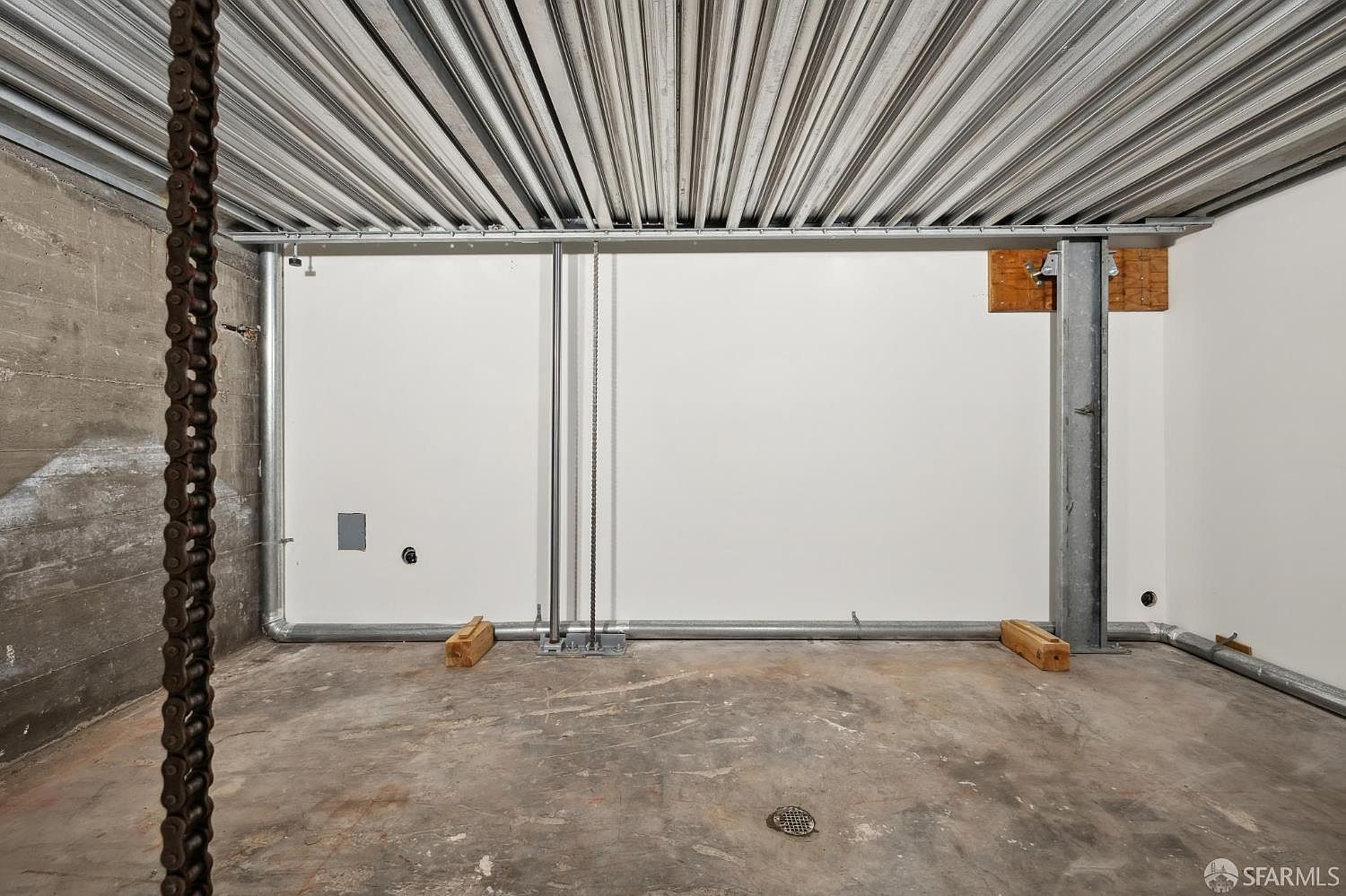This utilitarian basement space features exposed corrugated metal ceiling panels and raw concrete walls, suggesting an industrial or unfinished aesthetic. A prominent steel support column stands against the back wall, while exposed piping runs along the perimeter near the floor. The concrete floor includes a central drain, and the room is illuminated by overhead lighting, creating a stark, functional environment.