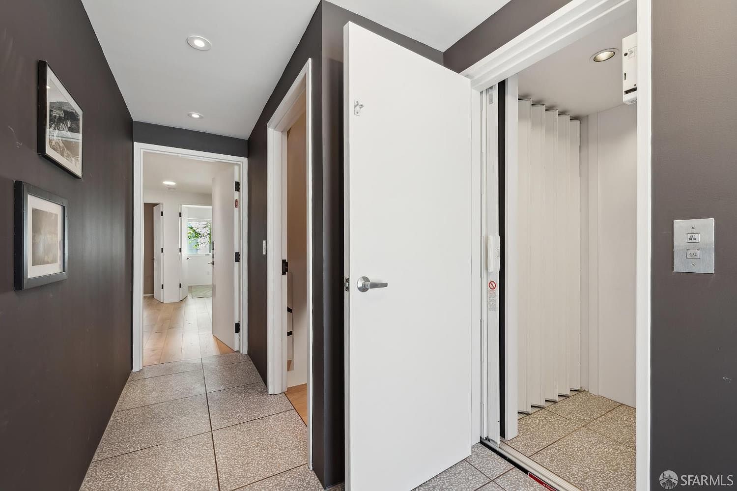 This interior hallway features dark-painted walls and light-colored tile flooring, creating a high-contrast, modern aesthetic. The space includes a white door leading to a secondary room, a view into a bright hallway beyond, and a private residential elevator with a folding door. The perspective is from the hallway looking toward the elevator, highlighting the functional layout and clean lines of the home.