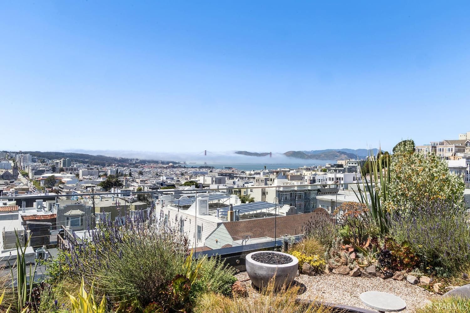 This elevated outdoor terrace offers a panoramic view of the San Francisco cityscape, including the iconic Golden Gate Bridge partially shrouded in fog. The space features a modern glass railing, a decorative stone fire pit, and a landscaped garden area with various succulents and grasses. The perspective is from a high vantage point, capturing the expansive urban horizon and the serene, private atmosphere of the rooftop deck.