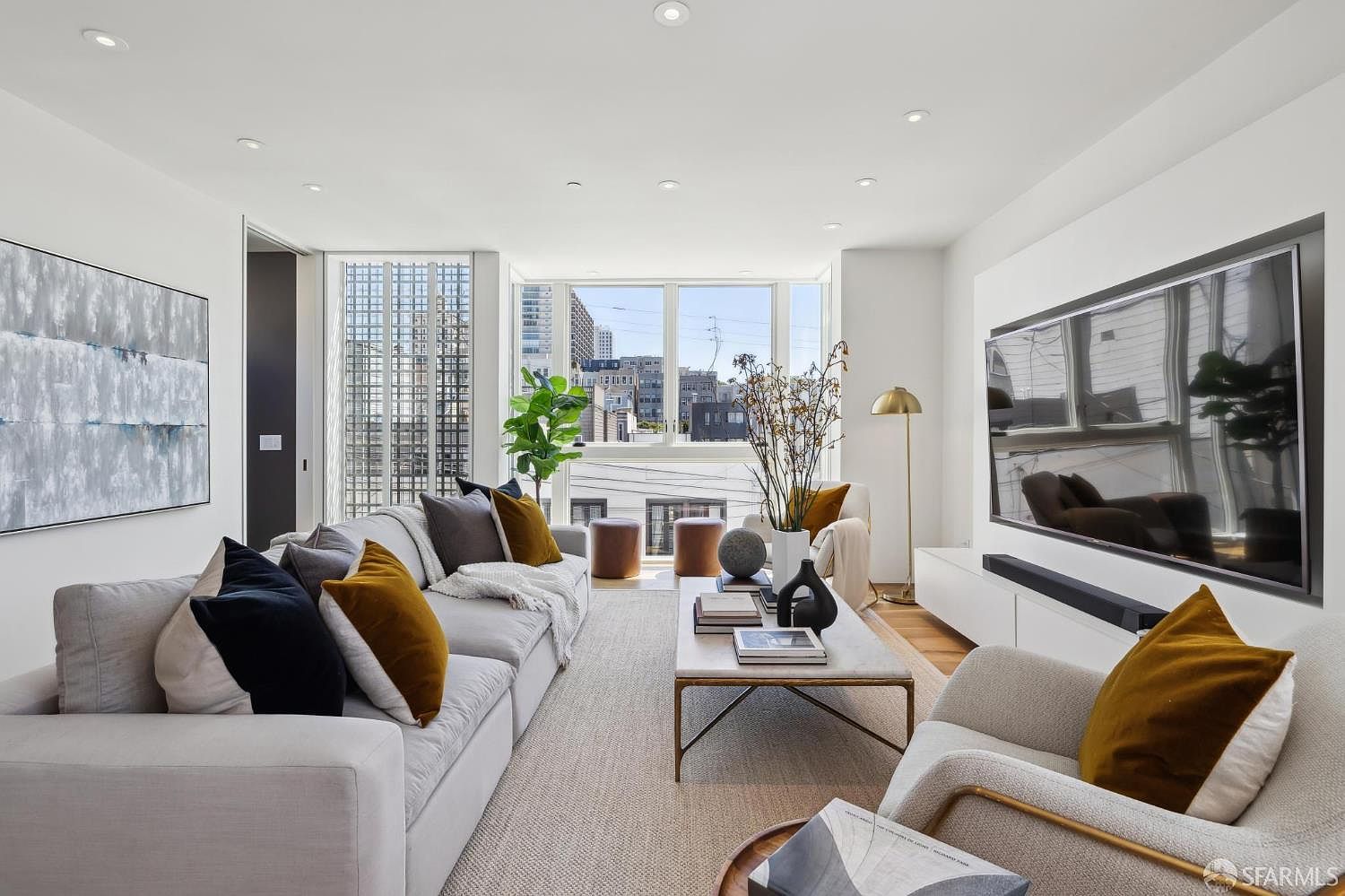 This bright and contemporary living room features a large, light-colored sectional sofa accented with dark and mustard-toned throw pillows. A central stone-topped coffee table sits on a textured area rug, while a large television is mounted on the wall opposite the seating area. Large windows at the far end of the room provide ample natural light and a view of the surrounding urban architecture.