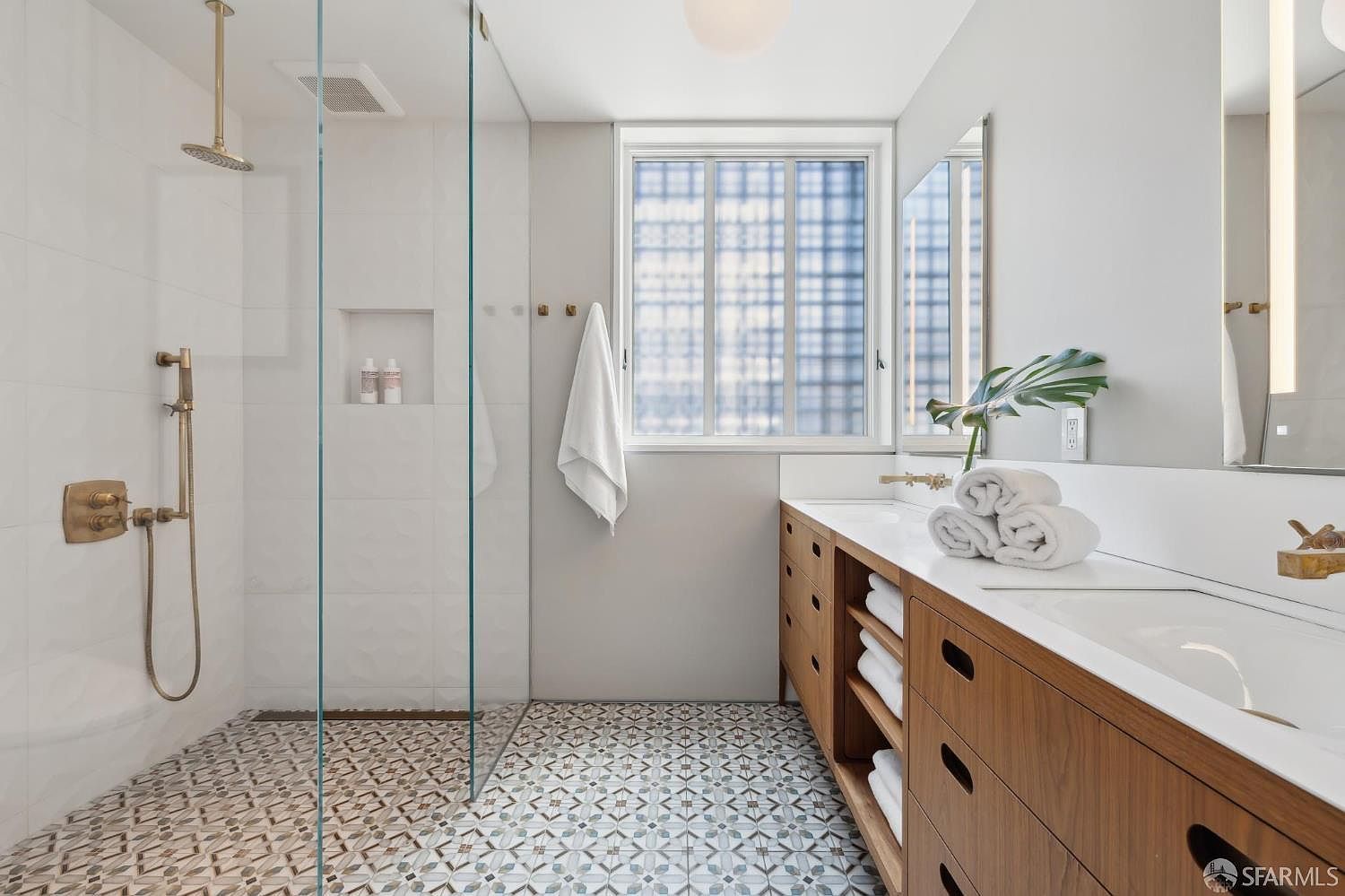 This modern bathroom features a spacious walk-in shower with a glass partition, a rainfall showerhead, and a built-in wall niche. A long vanity with light wood cabinetry and a white countertop provides ample storage, complemented by patterned floor tiles that add visual interest. The room is bright and airy, with a large window above the vanity area allowing natural light to fill the space.