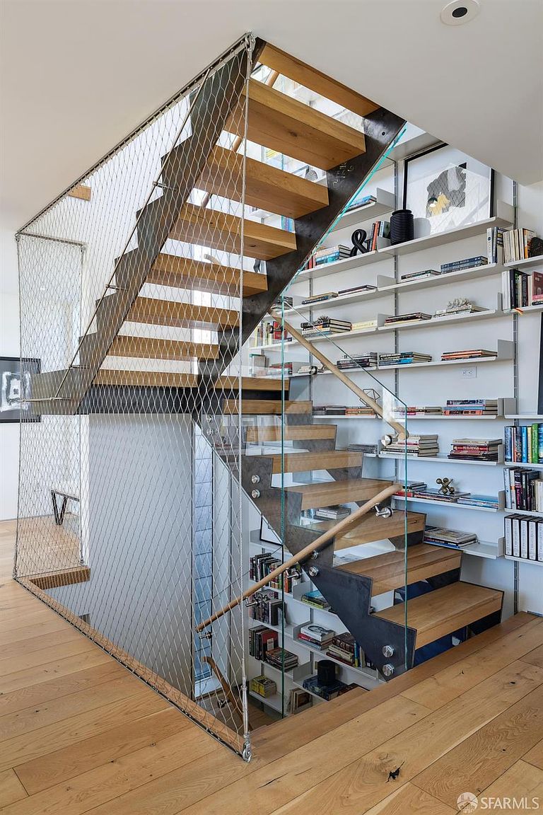 This modern staircase features light wood treads, a dark metal frame, and a unique wire mesh safety railing that creates an industrial-chic aesthetic. The adjacent wall is lined with extensive built-in shelving, providing ample space for books and decor, while a glass panel railing adds a sleek, transparent element to the design. The perspective captures the staircase from the upper landing, highlighting the open, airy feel and the clever integration of storage into the vertical space.