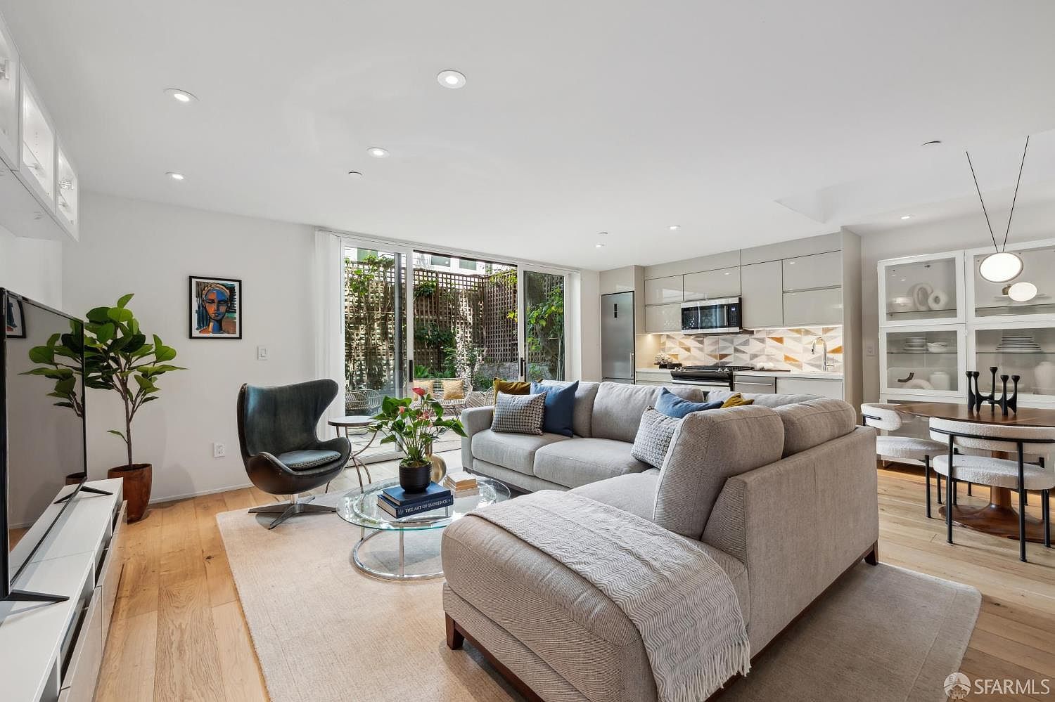 This open-concept living area features a light-toned sectional sofa, a modern egg-style accent chair, and a glass-topped coffee table set upon a neutral area rug. The space flows seamlessly into a contemporary kitchen with sleek cabinetry and a geometric-patterned backsplash, while large sliding glass doors provide a view of a private, lush outdoor patio. The room is bright and airy, characterized by light wood flooring and a clean, minimalist aesthetic.