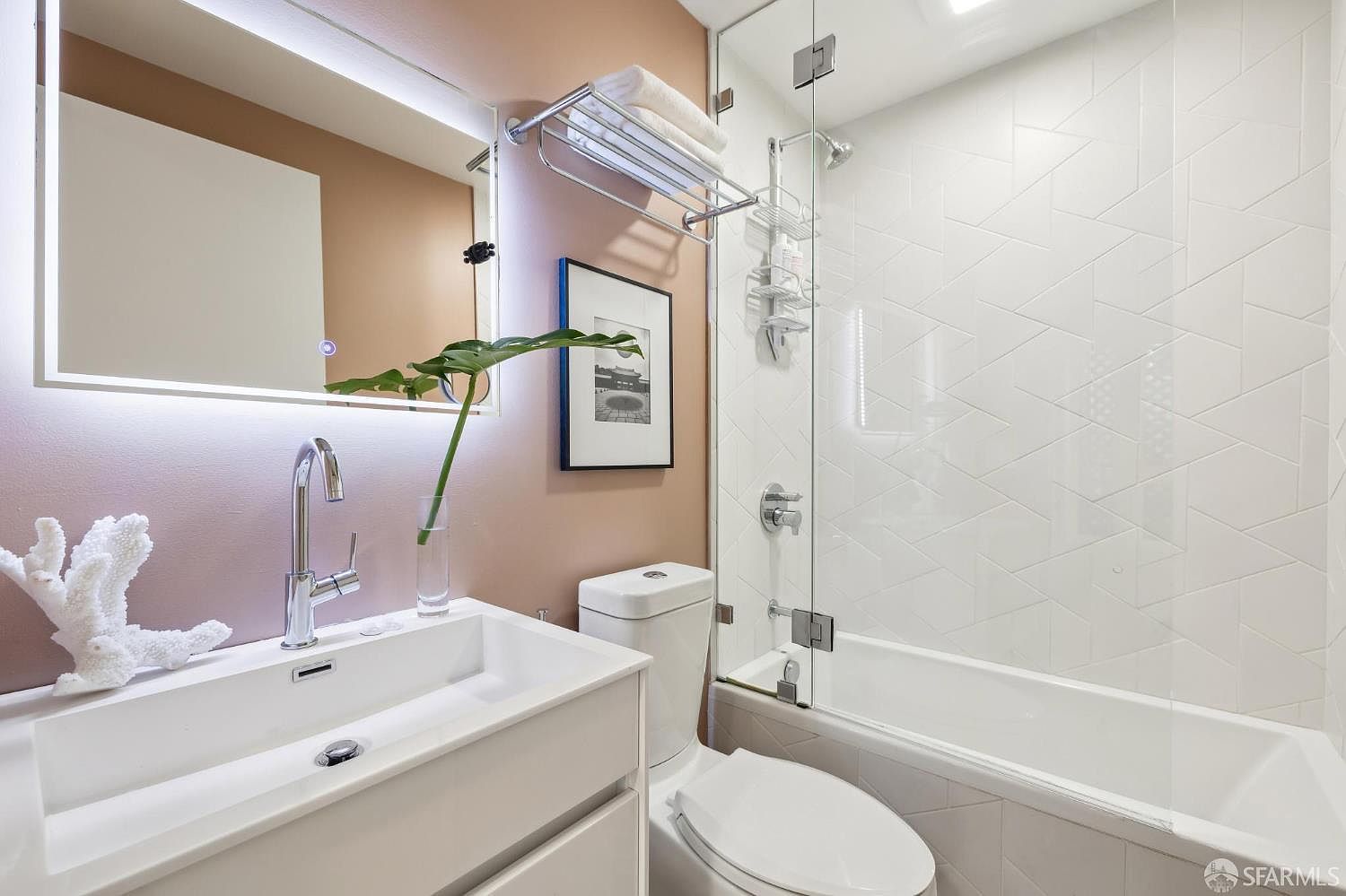 This modern bathroom features a sleek, wall-mounted vanity with a rectangular sink and a contemporary faucet, complemented by a large, backlit mirror. The space includes a shower-tub combination enclosed by a glass panel, finished with white geometric patterned wall tiles. The room is painted in a soft, warm neutral tone, creating a clean and inviting atmosphere.