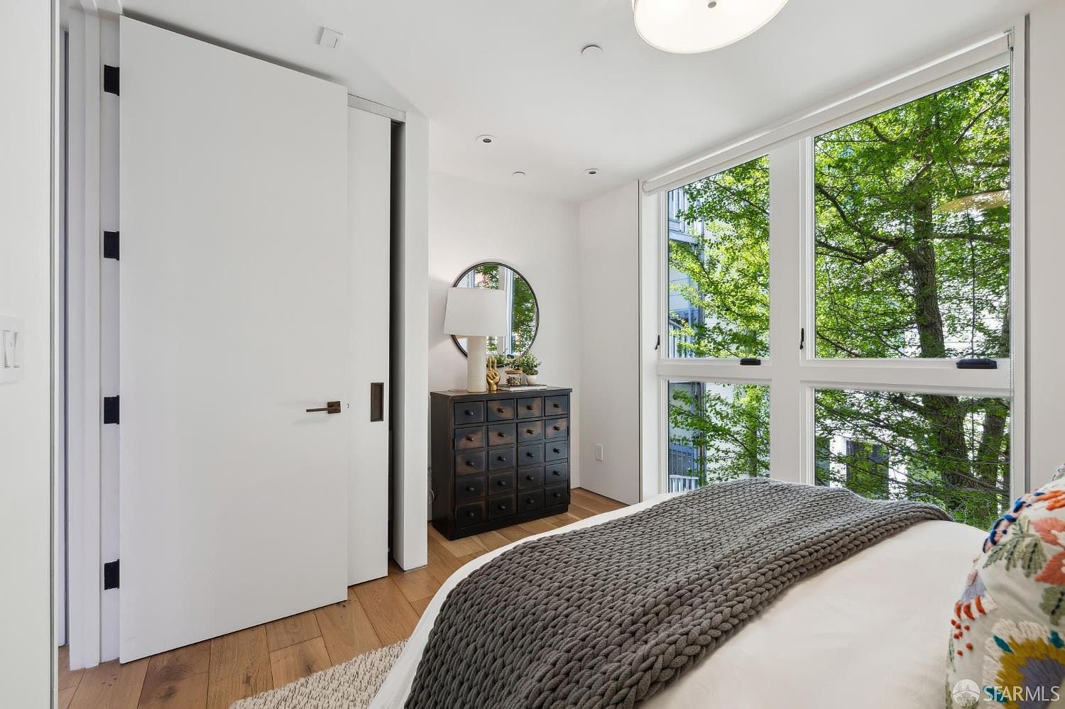 This bright and modern bedroom features a large bed with a textured gray knit throw blanket in the foreground. To the side, a dark wood chest of drawers with numerous small drawers sits beneath a round mirror and a table lamp, while a sliding white door provides access to an adjacent space. Large windows offer a serene, elevated view of lush green trees, filling the room with natural light.