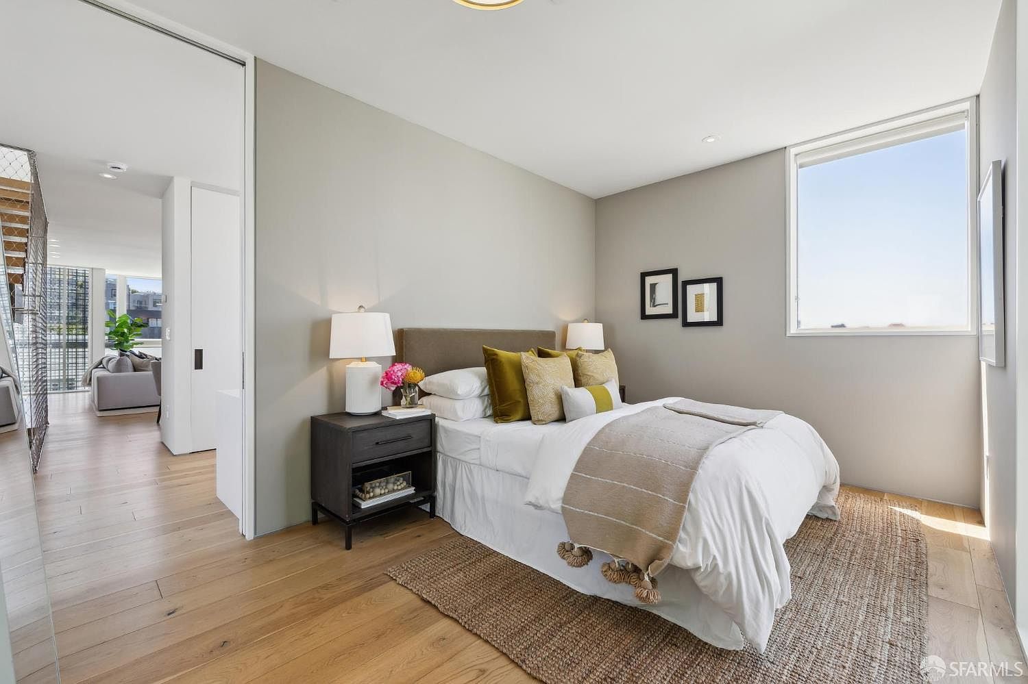 This bright and contemporary guest bedroom features light wood flooring and a neutral color palette, accented by mustard-toned throw pillows and a textured rug. A dark wood nightstand sits beside the bed, while a large window allows for ample natural light. The room offers a view into an adjacent living space, creating an open and airy feel throughout the home.