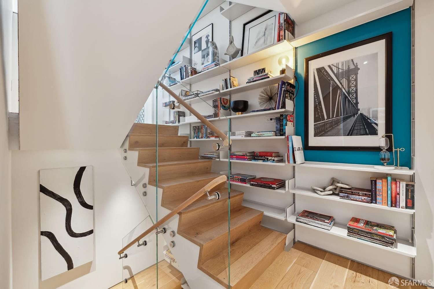 This modern staircase features light wood treads and a sleek glass railing, creating an open and airy aesthetic. A built-in shelving unit lines the wall, displaying books and decor against a vibrant teal accent wall, while a large framed black-and-white photograph adds a sophisticated touch. The perspective captures the transition between levels, highlighting the clean lines and contemporary design of the home.
