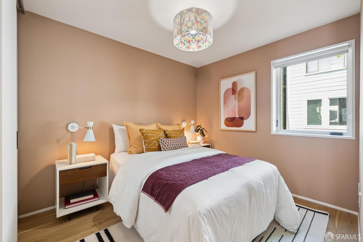 This cozy bedroom features warm, muted-tone walls and light wood flooring, creating a welcoming and serene atmosphere. The space is anchored by a bed dressed in white linens with a deep purple throw and decorative mustard-yellow pillows, complemented by a modern patterned drum pendant light. A large window allows natural light to fill the room, while a minimalist nightstand and abstract wall art add a touch of contemporary style.