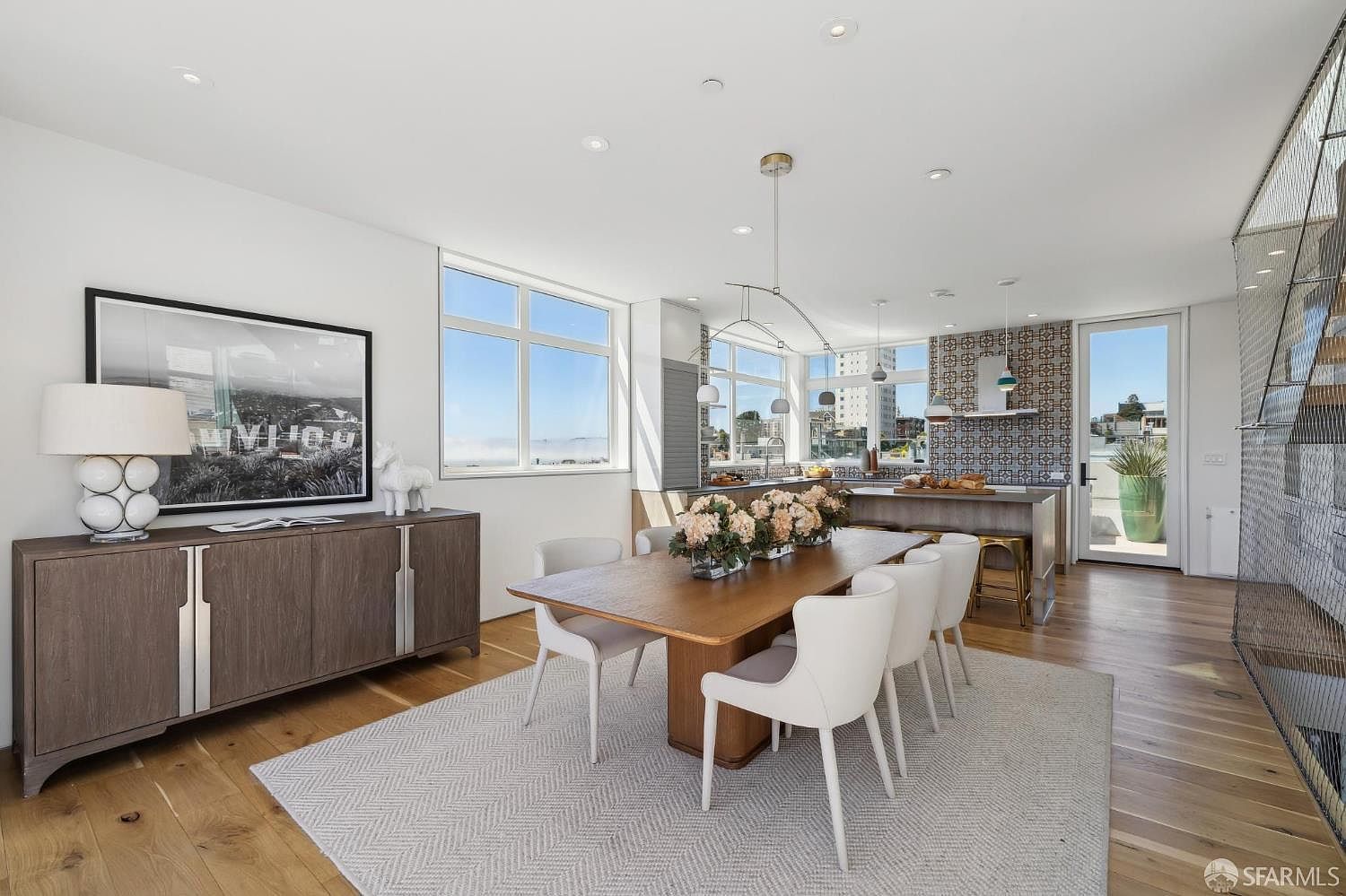 This bright and open-concept dining area features a large wooden table surrounded by modern white chairs, set atop a textured area rug. To the left, a long sideboard with a decorative lamp and framed artwork provides storage, while the background reveals a kitchen with a patterned tile backsplash and a breakfast bar. Large windows allow for abundant natural light, creating an airy and sophisticated atmosphere suitable for contemporary living.