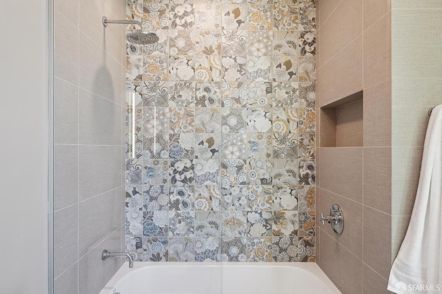 This bathroom features a shower-tub combination highlighted by a striking accent wall of patterned floral tiles in muted earth tones. The surrounding walls are finished in neutral, textured tiles, complemented by a built-in recessed niche for storage and polished chrome fixtures. A glass panel provides a clean, modern look to the bathing area, creating a sophisticated and serene atmosphere.