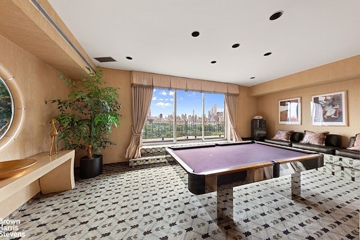 This spacious game room features a prominent purple-felt pool table as its centerpiece, set against a backdrop of large windows that offer a panoramic view of the city skyline and park. The room is finished with light wood-toned wall paneling, a patterned carpet, and recessed ceiling lighting, creating a sophisticated entertainment space. A large potted plant adds a touch of greenery, while a leather sofa provides comfortable seating along the side wall.