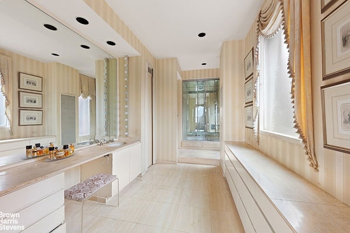This elegant primary bathroom features a long vanity with a large mirror and integrated lighting, complemented by a matching built-in storage console along the opposite wall. The space is finished with light-toned striped wallpaper, neutral tile flooring, and a glass-enclosed shower area at the far end. The room is accented with framed artwork and soft, draped window treatments, creating a sophisticated and cohesive aesthetic.
