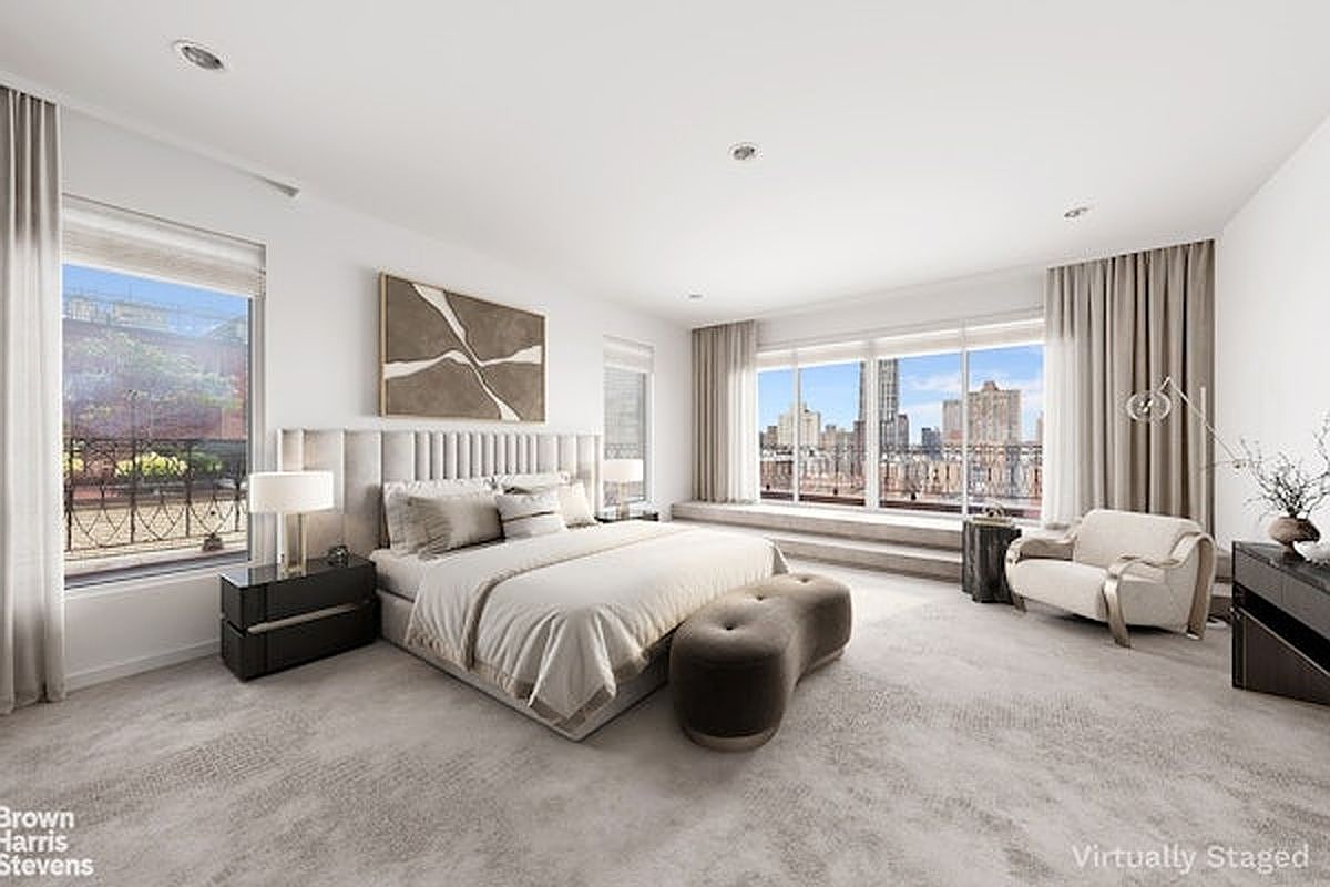 This spacious primary bedroom features a neutral color palette, plush carpeting, and a large upholstered headboard centered against a white wall. Large windows provide expansive city views and allow for abundant natural light, while a curved ottoman sits at the foot of the bed. The room is furnished with a modern armchair and sleek bedside tables, creating a sophisticated and serene atmosphere.