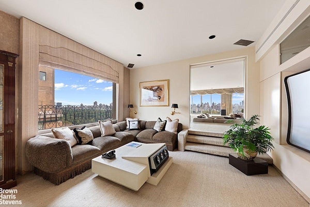 This sophisticated living room features a large, plush sectional sofa arranged around a unique, tiered white coffee table. A expansive window provides a panoramic view of a city park, while an adjacent elevated area with a mirror wall adds depth and a sense of openness to the space. The room is decorated in a neutral, monochromatic palette with soft carpeting and warm lighting, creating an elegant and inviting atmosphere.