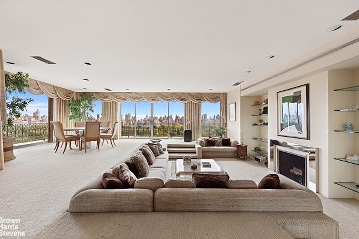 This expansive, open-concept living area features a neutral color palette and plush carpeting, centered around a large, L-shaped sectional sofa. A fireplace with a sleek, metallic surround serves as a focal point, while a wall of large windows provides panoramic views of the city skyline and park. The space flows seamlessly into a dining area, creating an airy and sophisticated atmosphere perfect for entertaining.