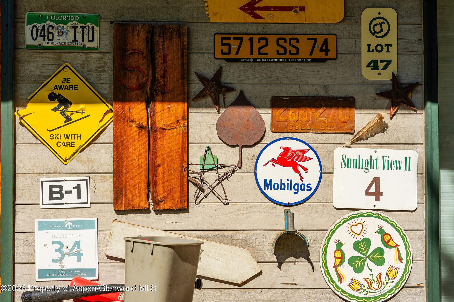 This image captures a rustic, weathered wood-paneled wall adorned with a eclectic collection of vintage signs, metal art, and decorative plaques. The display includes a Mobilgas sign, a Colorado license plate, and various directional and informational markers, creating a nostalgic, Americana-inspired aesthetic. The composition is a close-up, static shot that highlights the textured wood grain and the patina of the various metal objects.