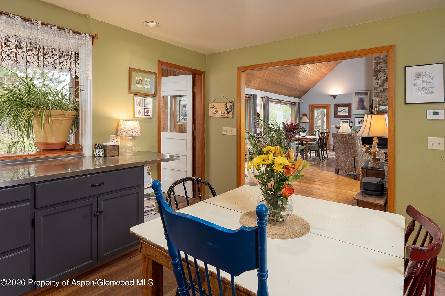 This cozy dining area features a white-painted wooden table centered in the room, surrounded by chairs with blue and red accents. The space opens into an adjacent living area, which showcases a stone-clad chimney breast and vaulted wood-paneled ceilings. A stainless steel counter with dark cabinetry sits to the left, while a large window with a potted plant provides natural light, creating a warm and inviting atmosphere.