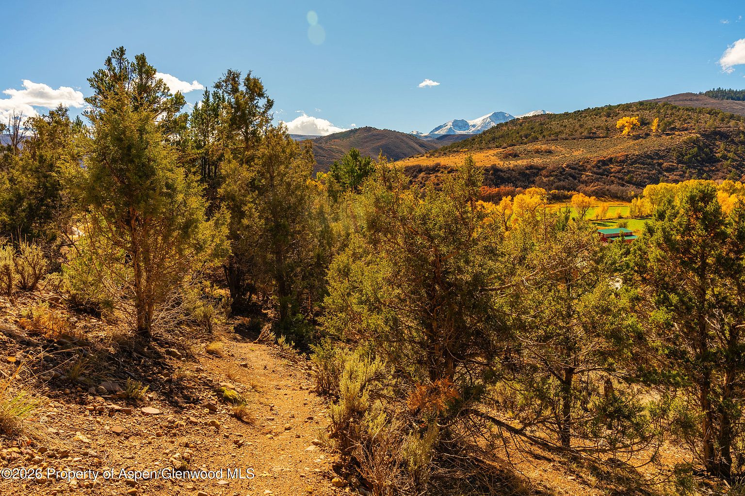 This scenic landscape view captures a rugged, sun-drenched dirt path winding through a grove of juniper and pine trees. In the distance, a vibrant valley floor with golden autumn foliage leads to a small structure, all set against a backdrop of majestic, snow-capped mountain peaks under a clear blue sky. The perspective offers a wide, elevated vantage point that emphasizes the expansive natural beauty and serene atmosphere of the property.