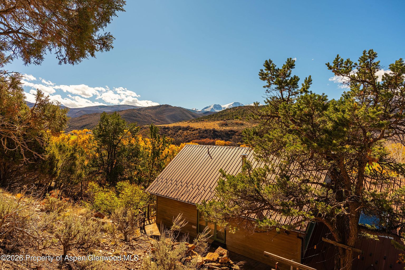 This elevated, high-angle shot captures a rustic cabin nestled within a dense, wooded landscape during the autumn season. The home features a simple gabled roof and wood siding, surrounded by vibrant yellow foliage and evergreen trees, with a dramatic mountain range visible in the distance under a clear blue sky. The perspective emphasizes the secluded, natural setting of the property, highlighting its integration into the rugged terrain.