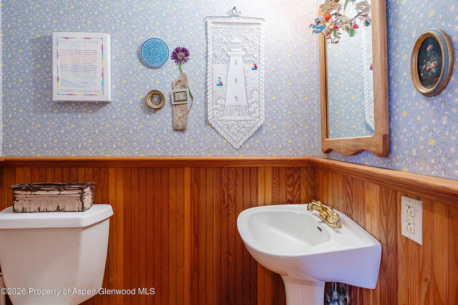 This powder room features a classic aesthetic with floral wallpaper above a wainscoting of warm-toned wood paneling. A white pedestal sink with gold-toned hardware sits against the wall, complemented by a mirror adorned with decorative accents and various wall-mounted art pieces. The space is compact and functional, offering a cozy and traditional atmosphere.