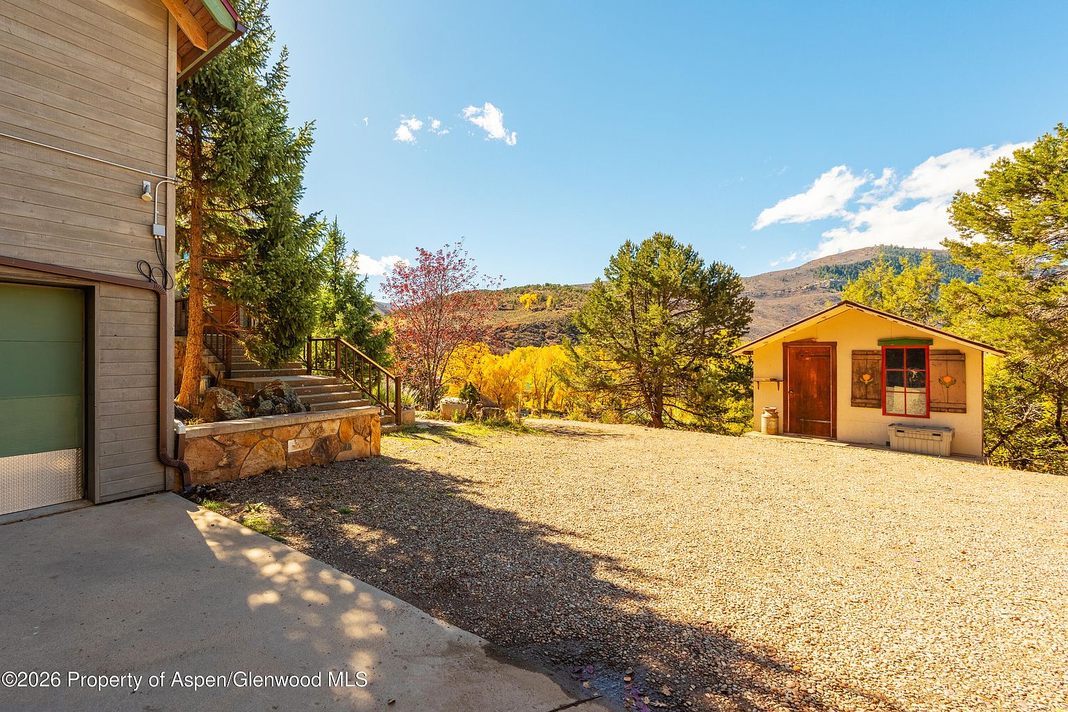 This outdoor scene captures a gravel driveway leading toward a small, rustic shed with a red-trimmed window and wooden door. To the left, a stone retaining wall supports a set of stairs ascending toward a house, framed by mature evergreen trees and vibrant autumn foliage in the background. The perspective is a wide-angle shot from the driveway, emphasizing the expansive mountain landscape and the peaceful, rural setting.