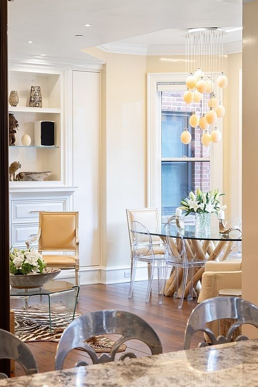 This is an interior shot of a dining room featuring a glass-topped table with a unique wooden base, surrounded by clear acrylic chairs. A modern chandelier hangs above the table, and built-in shelving with decorative items is visible in the background. The room has a bright and airy feel, enhanced by the natural light coming through the window.