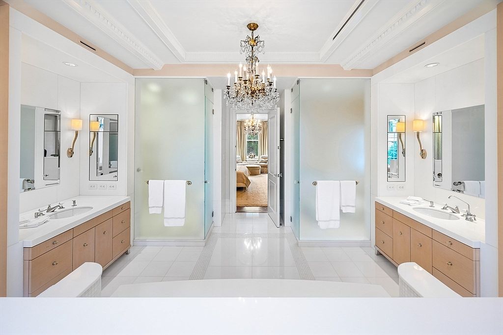 This is a luxurious primary bathroom featuring dual vanities with light wood cabinetry and white countertops. Frosted glass doors lead to a bedroom, and a crystal chandelier hangs from the ceiling, adding a touch of elegance. The white tile flooring and walls create a bright and clean atmosphere.